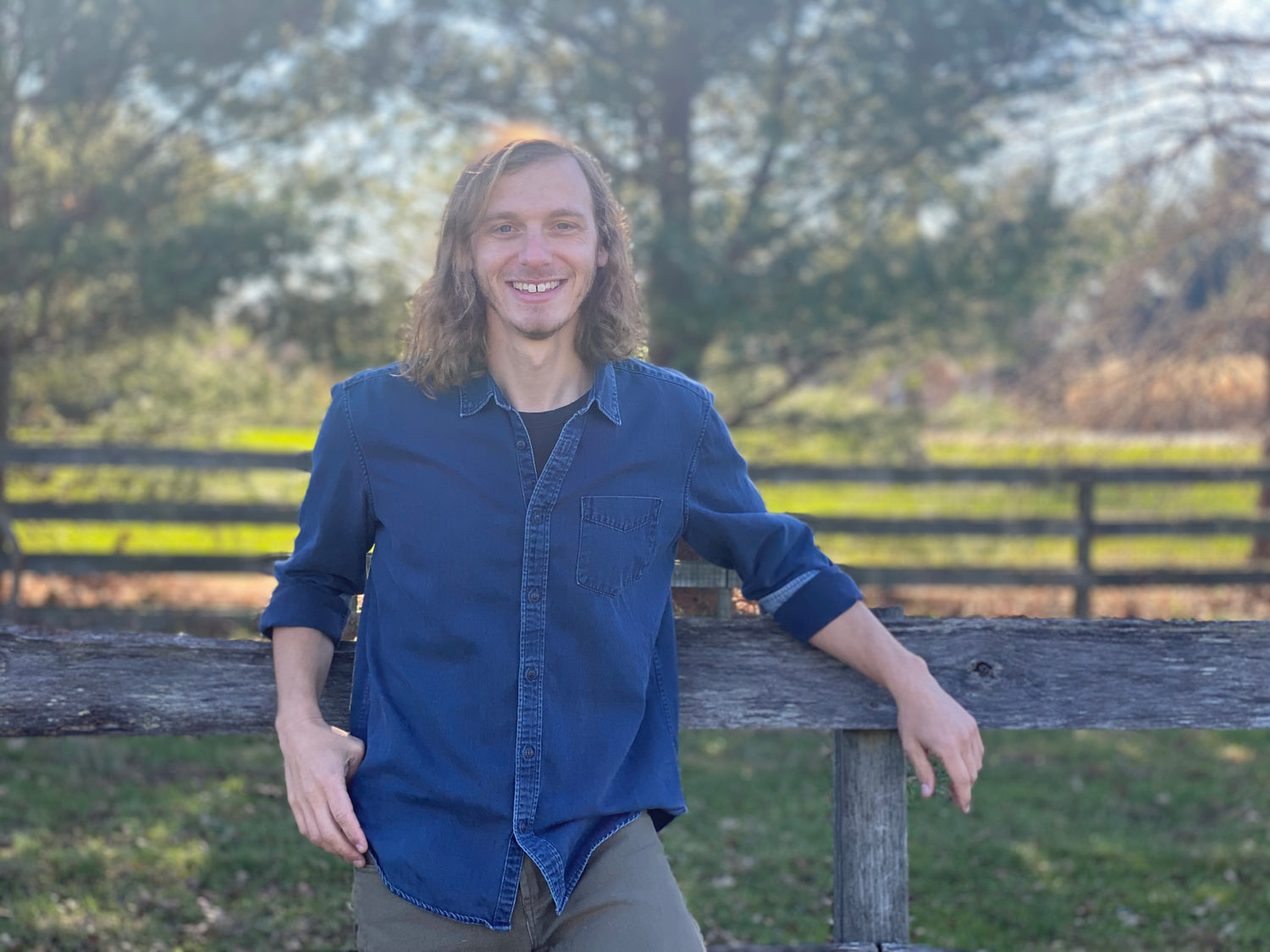 Carpenter Joe Hedglin stands against a wooden fence, casually leaning up against it. He has long, wavy blond hair and is wearing a dark blue button down shirt.